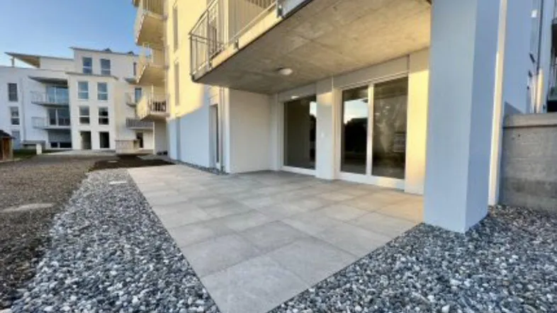 Eine moderne Terrasse im Erdgeschoss eines Neubaus, umgeben von Kies und mit Blick auf weitere Gebäude.