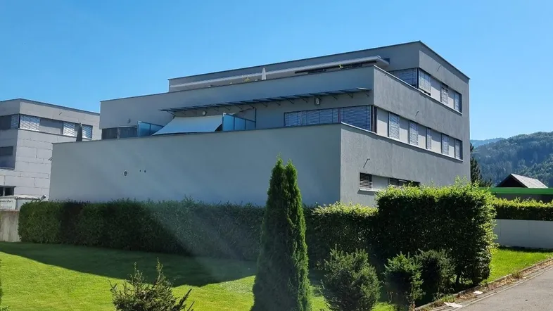 Modernes Gebäude mit grauer Fassade, umgeben von gepflegtem Rasen und Hecken unter blauem Himmel.