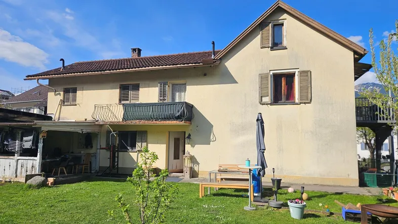 Rückseite eines Hauses mit grünem Garten, Balkon und Sitzbereich unter blauem Himmel.