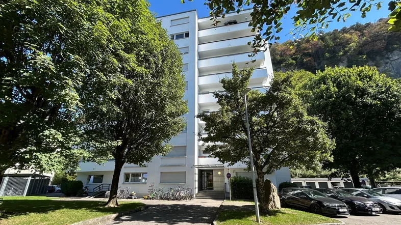 Modernes Mehrfamilienhaus mit Balkonen, umgeben von Bäumen und Parkplätzen, vor einer Bergkulisse.