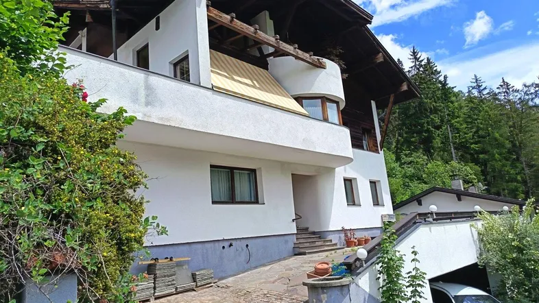 Mehrstöckiges Haus mit weißer Fassade, Holzbalkonen und einer Einfahrt mit Garage, umgeben von Bäumen.