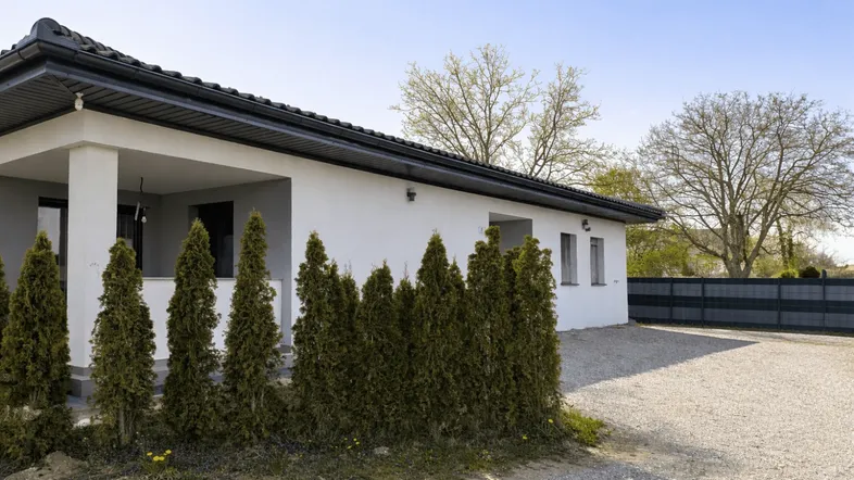 Modernes Einfamilienhaus mit weißer Fassade, Kiesauffahrt und grüner Hecke unter blauem Himmel.