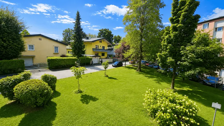 Grüne Rasenfläche mit Büschen und Bäumen, umgeben von Wohnhäusern unter blauem Himmel.