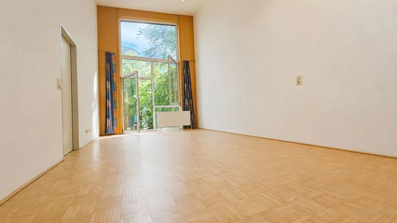 Helles, leeres Zimmer mit Parkettboden und großen Fenstern mit Blick ins Grüne.