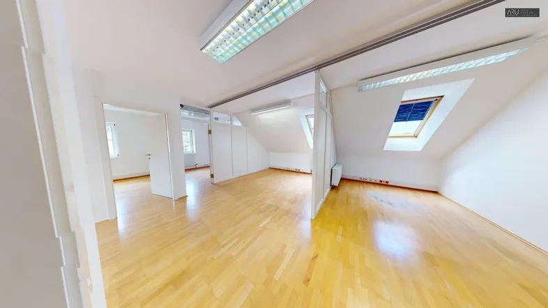 Heller Innenraum mit Holzfußboden, weißen Wänden und Dachschrägen, ausgestattet mit Deckenleuchten und Dachfenster.