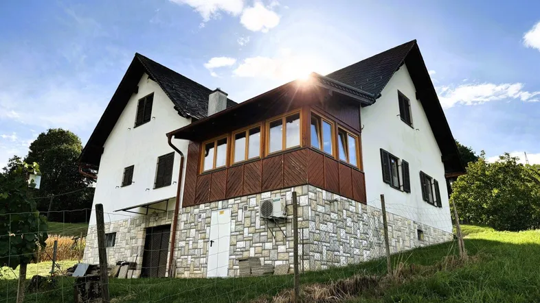 Traditionelles Haus mit Steinsockel und Holzanbau, umgeben von grüner Landschaft unter strahlendem Himmel.