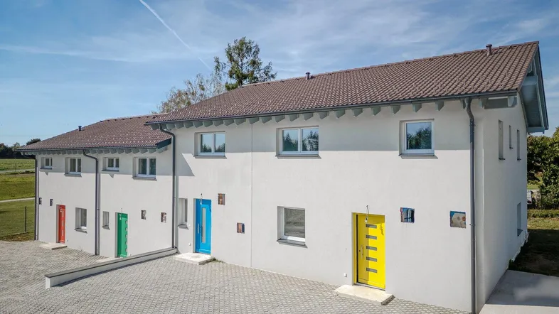 Moderne Reihenhäuser mit farbigen Eingangstüren und gepflastertem Vorplatz unter blauem Himmel.
