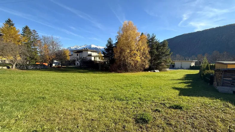 Weitläufige Grünfläche mit Blick auf die umliegende Berglandschaft und benachbarte Häuser unter blauem Himmel.
