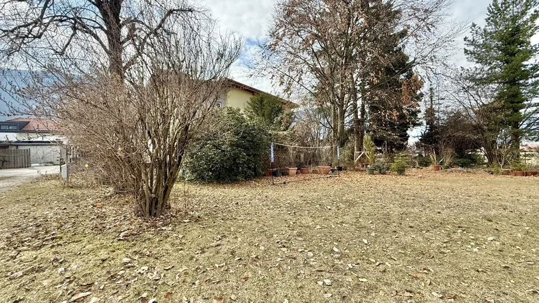 Großer Gartenbereich mit Rasenfläche und Bäumen, im Hintergrund ein Wohnhaus unter bewölktem Himmel.