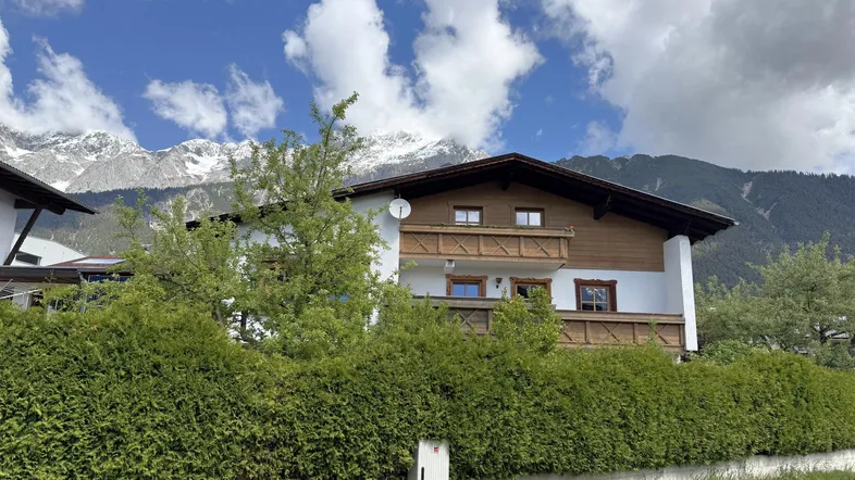 Traditionelles Haus mit Holzbalkon und weißer Fassade vor einer beeindruckenden Bergkulisse.