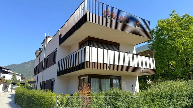 Modernes Mehrfamilienhaus mit mehreren Balkonen und gepflegter Hecke in bergiger Umgebung.