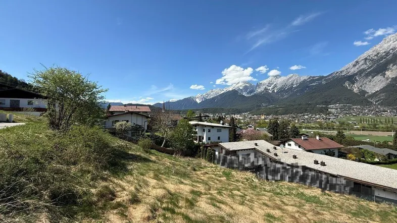 Panoramablick auf eine Wohnsiedlung mit grünen Hügeln und schneebedeckten Bergen im Hintergrund unter blauem Himmel.