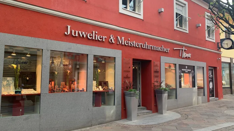 Rote Geschäftsfassade eines Juweliers mit großen Schaufenstern, Eingang und dekorativen Pflanzen am Bürgersteig.