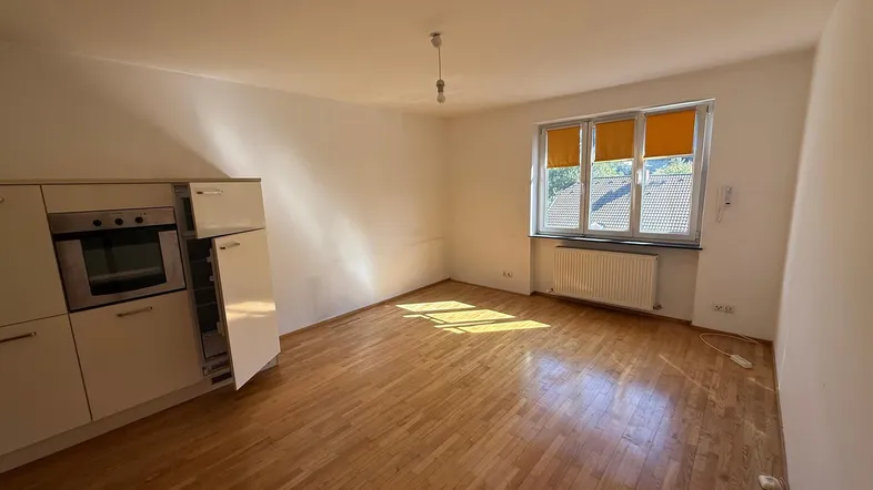 Helles Zimmer mit Holzboden, weißen Wänden und einer kleinen Küchenzeile. Sonnenlicht fällt durch das Fenster.