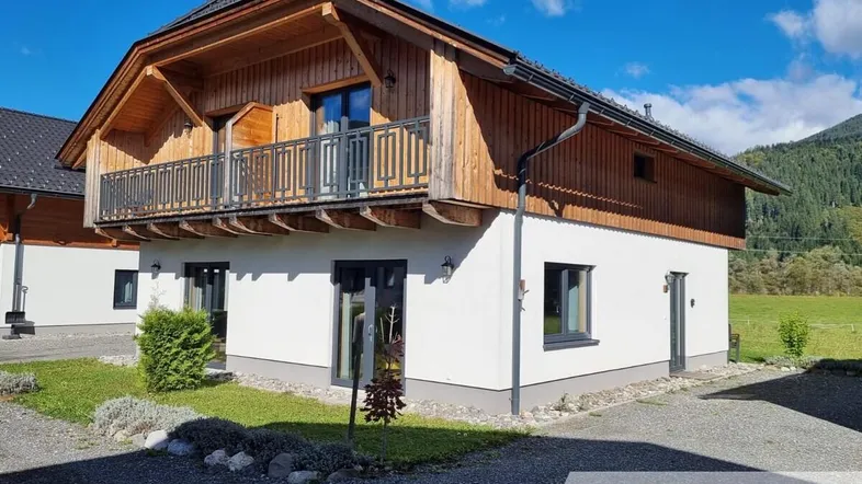 Charmantes Holzhaus mit weißer Fassade und Balkon in ländlicher Umgebung unter blauem Himmel.