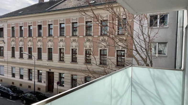 Blick vom modernen Balkon mit Glasbrüstung auf die gegenüberliegende historische Fassade und die Straße.