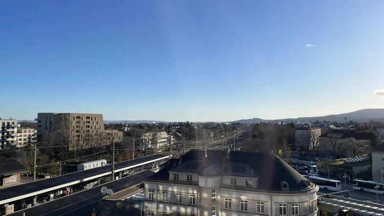 Panoramablick über die Stadt mit Bahnhof, Gebäuden und Bergen im Hintergrund unter blauem Himmel.