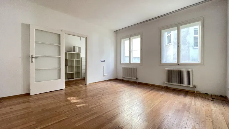 Helles Wohnzimmer mit Holzboden und zwei Fenstern, die viel Tageslicht hereinlassen.