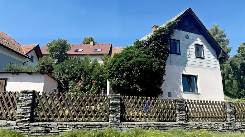 Hausfassade mit Kletterpflanzen und einem rustikalen Holzzaun, umgeben von grüner Natur.