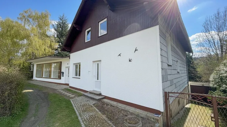 Vorderansicht eines Hauses mit weißer Fassade, Holzelementen und gepflastertem Weg unter strahlendem Sonnenschein.
