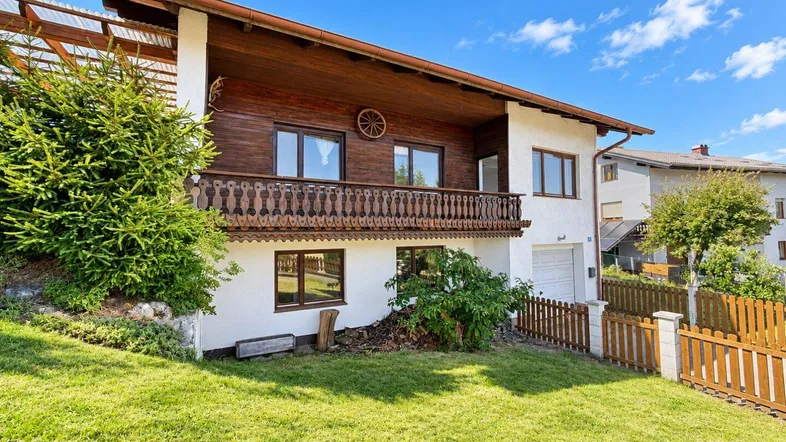 Traditionelles Haus mit Holzbalkon und gepflegtem Vorgarten unter blauem Himmel.