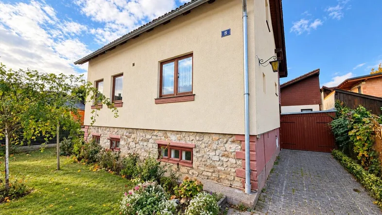 Seitenansicht des Hauses mit gepflegtem Vorgarten, Steinmauer und gepflasterter Einfahrt unter blauem Himmel.