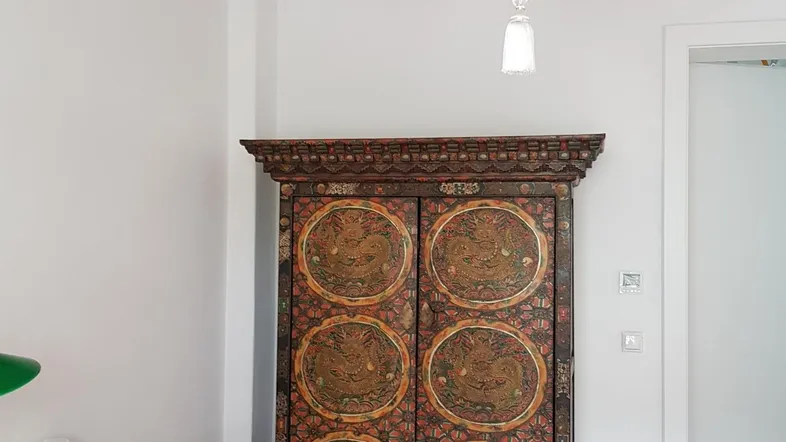 Helles Büro mit Holztisch, Stuhl, einem kunstvoll verzierten Schrank und sichtbaren Deckenbalken.