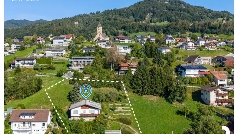 Luftaufnahme eines Einfamilienhauses auf einem grünen Hügel mit umliegender Bebauung und Bergpanorama.