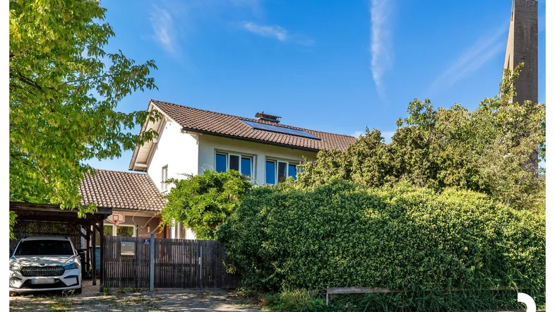 Weißes Einfamilienhaus mit Carport, umgeben von viel Grün und einem Kirchturm im Hintergrund.