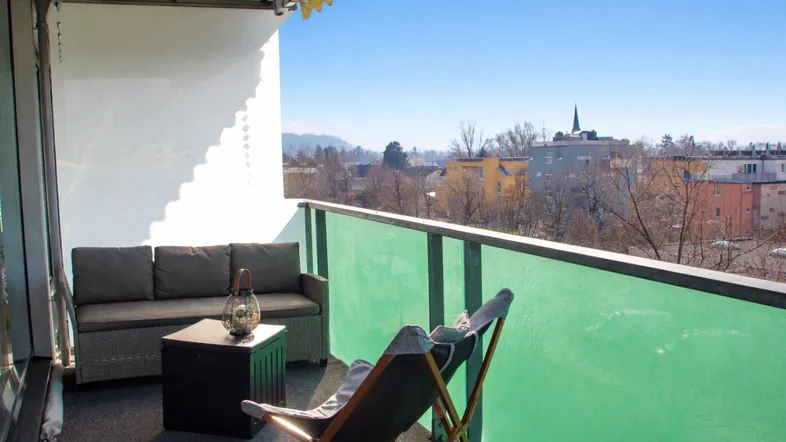 Möblierter Balkon mit grüner Brüstung und weitem Blick über die Stadt und Berge.