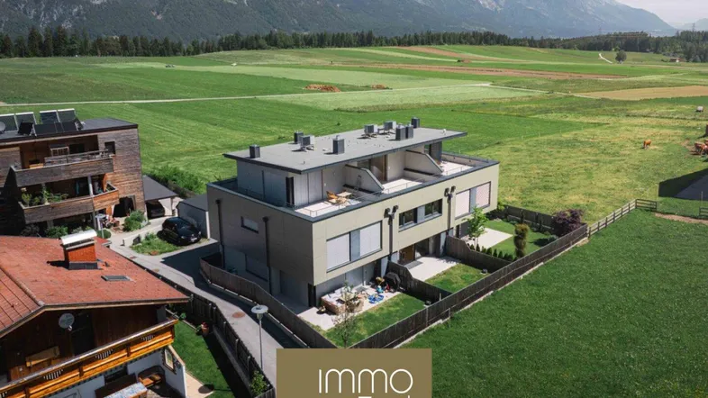 Moderne Wohnanlage mit Flachdach und großen Fenstern, umgeben von grünen Feldern und Bergen.