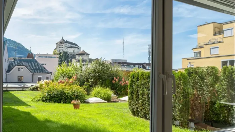 Blick aus einem Fenster auf einen begrünten Dachgarten mit Schloss im Hintergrund und blauem Himmel.