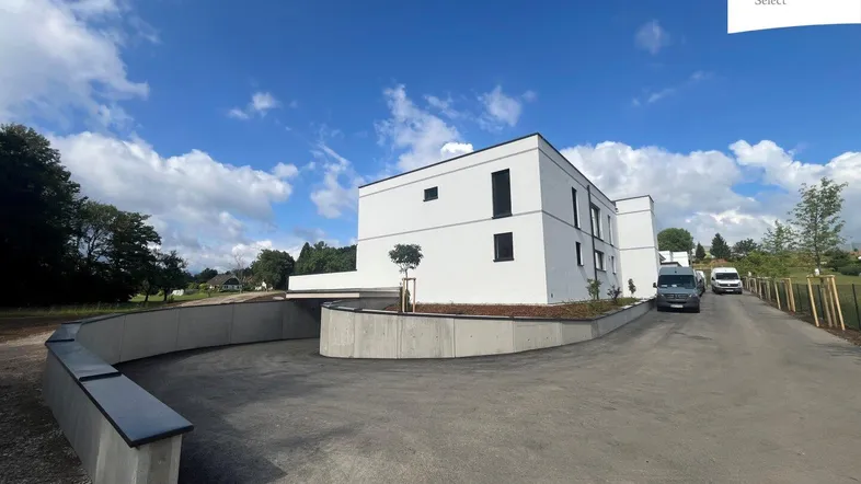 Modernes weißes Gebäude mit Flachdach und Zufahrt zu einer Tiefgarage unter blauem Himmel.