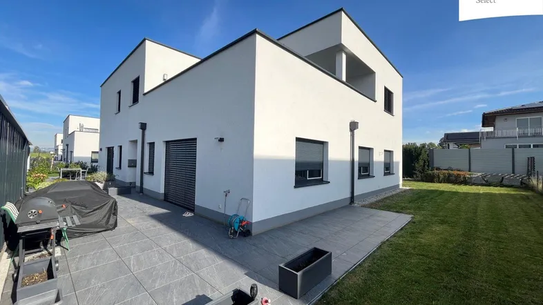 Modernes Einfamilienhaus mit weißer Fassade, gepflasterter Terrasse und Rasenfläche im Garten.