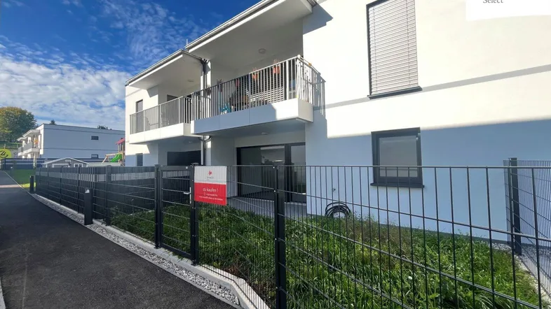 Modernes Mehrfamilienhaus mit Balkonen und eingezäuntem Gartenbereich unter blauem Himmel.