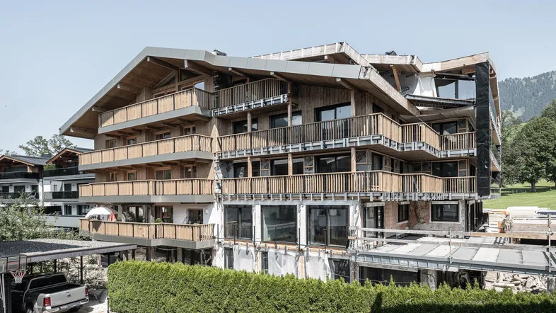 Großes Wohngebäude in Holzbauweise mit mehreren Balkonen, noch im Bau befindlich, vor alpiner Kulisse.