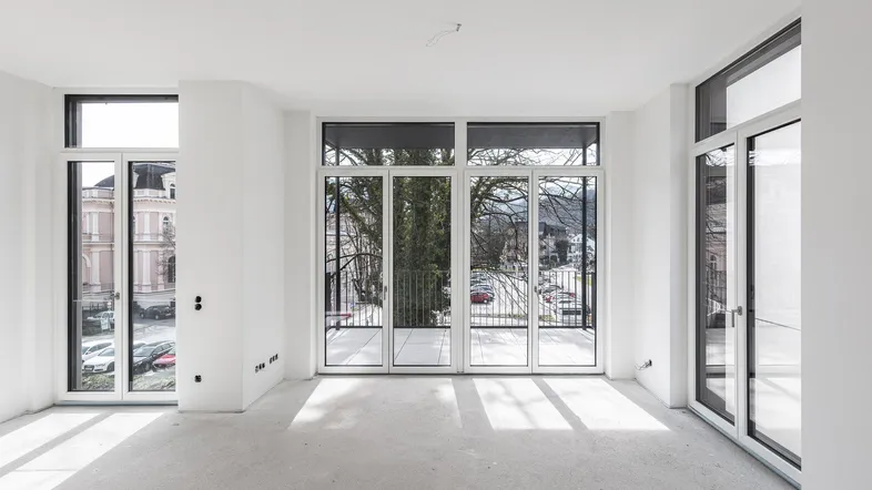 Heller, unmöblierter Raum mit großen Fenstern, hohen Decken und Zugang zu einem Balkon.