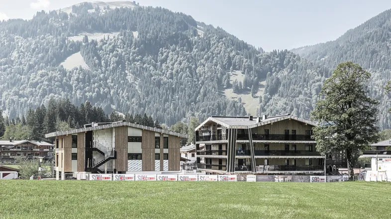 Zwei moderne Gebäude im Bau mit Holzfassaden und Bergblick in grüner Landschaft.
