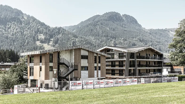 Zwei moderne Gebäude im Bau mit Holzfassaden und Blick auf die umliegenden Berge.