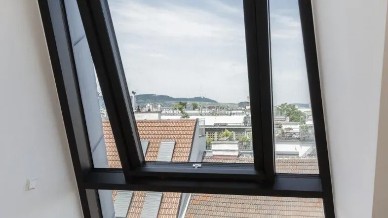 Helles Zimmer im Dachgeschoss mit schrägem Fenster und Blick über die Dächer der Stadt.