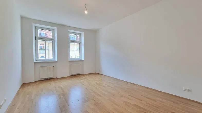 Helles, leeres Zimmer mit Parkettboden und zwei Fenstern, bereit zur individuellen Gestaltung.