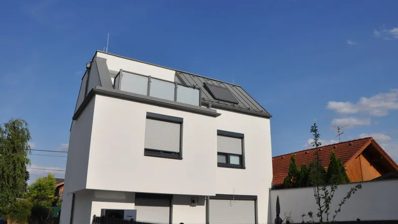 Modernes Wohnhaus mit weißer Fassade, grauem Dach und kleinem Balkon unter blauem Himmel.