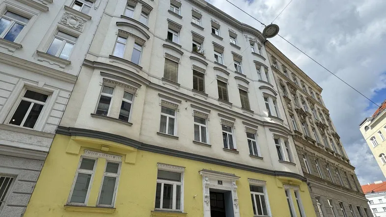 Historische Fassade eines Altbaus mit weißer Oberseite und gelb gestrichenem Erdgeschoss unter bewölktem Himmel.