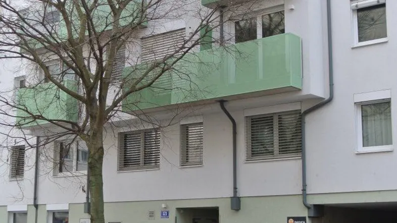 Mehrstöckiges Wohngebäude mit hellgrünen Balkonen und einem kahlen Baum davor an der Straße.