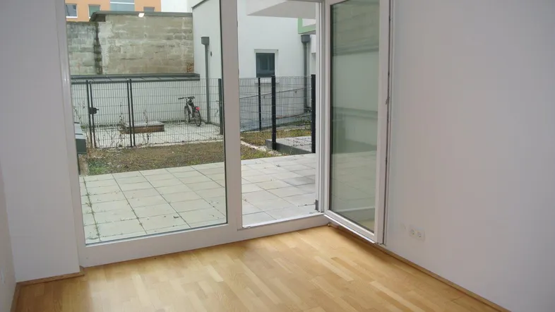 Helles Zimmer mit Parkettboden und direktem Zugang zu einer kleinen gefliesten Terrasse und Gartenbereich.