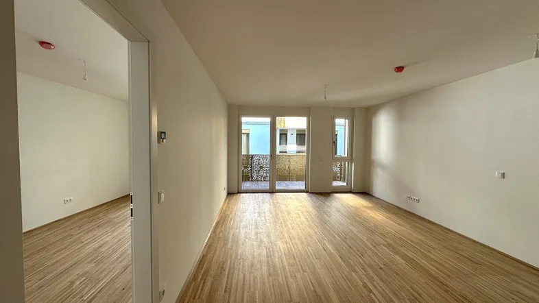Helles, leeres Wohnzimmer mit Holzboden und Zugang zu einem Balkon.