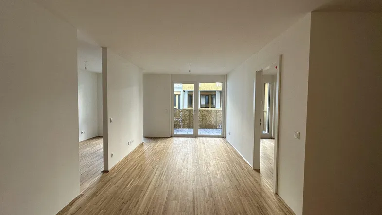 Geräumiges, leeres Wohnzimmer mit hellem Parkettboden und Zugang zu einem Balkon.