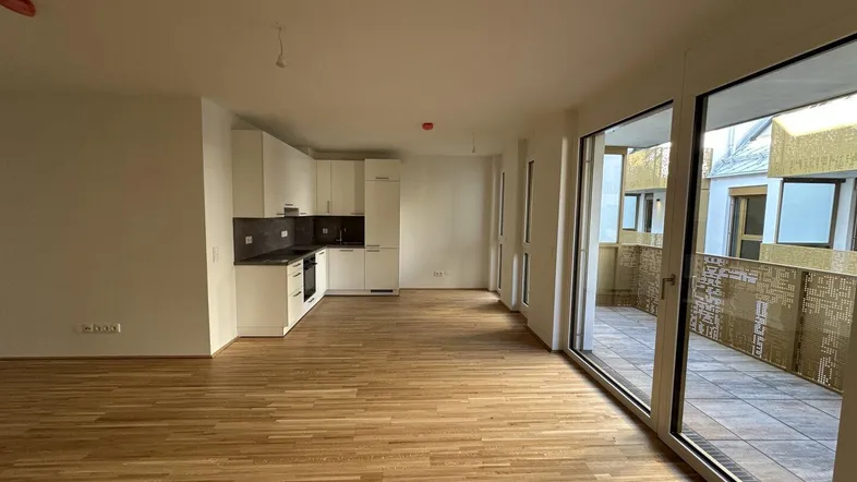 Helles Wohnzimmer mit integrierter Küche, Parkettboden und Zugang zu einem Balkon.