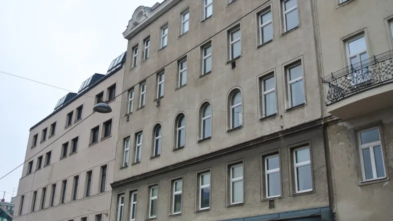 Graue Fassade eines Mehrfamilienhauses mit verschiedenen Fenstertypen und einem Balkon.