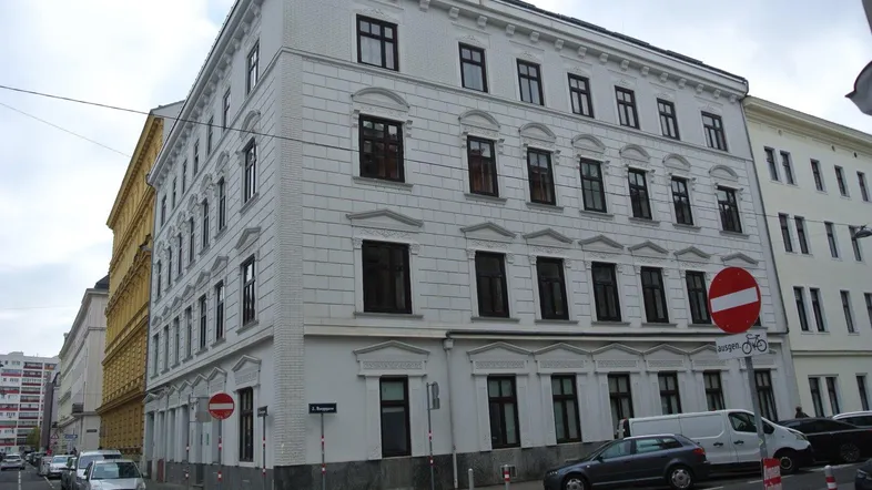 Weißes Eckgebäude mit vielen Fenstern und klassischer Architektur an einer Straße.